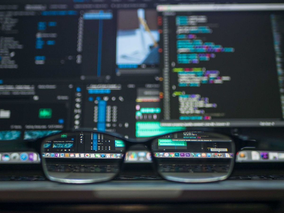 Glasses in front of monitors displaying code and a video editing interface